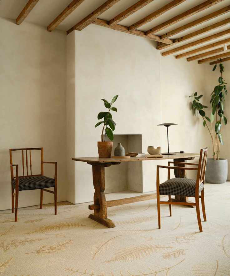 A light room with paster walls and an empty fireplace. A wooden desk with wooden arm chairs wither side of it. Carpeted floor with leaf patterns on it.