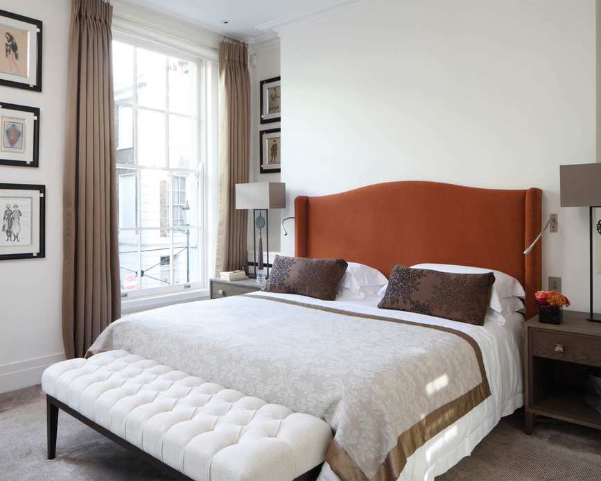 Bedroom showcasing a brown upholstered headboard, white bedding with brown cushions, and a cream ottoman with a black frame at the foot of the bed, with long brown curtains and dark wooden bedside tables.