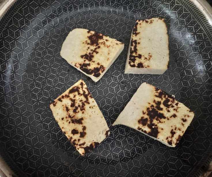 Four large chunks of tofu in a black lined frying pan, with char marks.