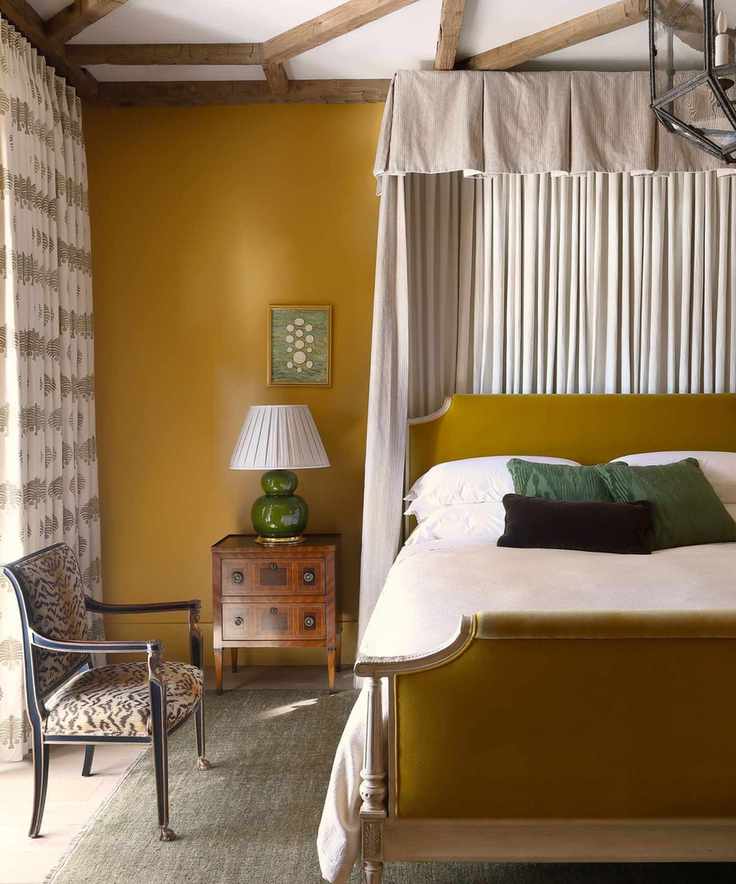 Bedroom with beamed ceiling, mustard velvet bed frame, mustard walls, antique bedside table and chair, and bed canopy