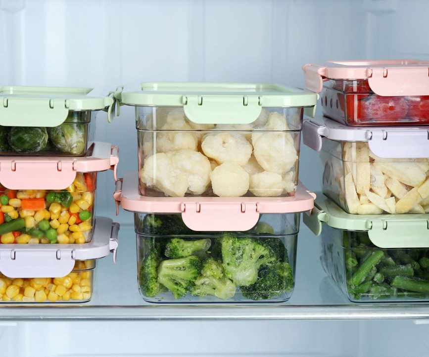 Glass food storage containers stacked with multi-colored lids in a fridge, filled with leftover vegetables
