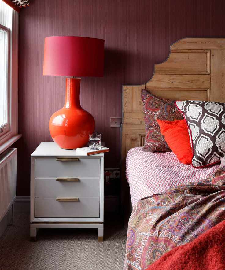 Red wallpaper in bedroom with eclectic red and geometric bedding, wooden headboard and large statement bedside lamp on chest of drawers, window to the left of the image, brown and white flecked carpet.