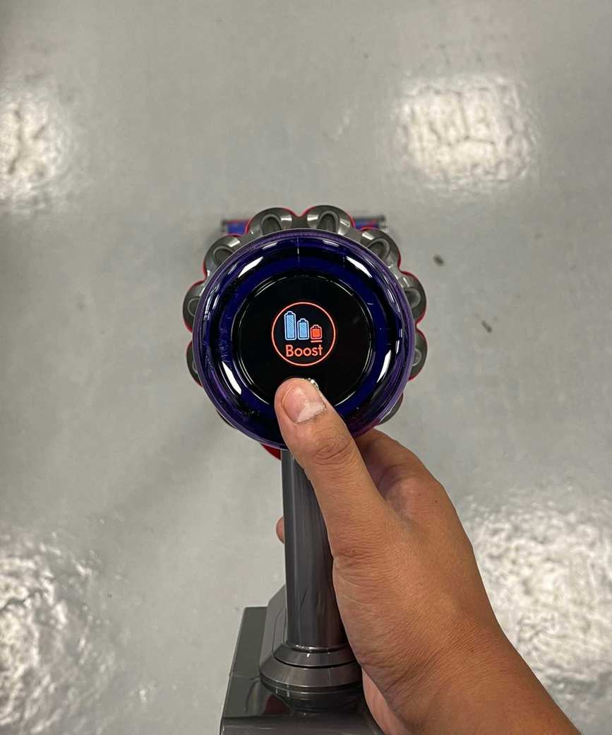 Dyson V11 LCD screen showing Boost mode, with Dan Fauzi's thumb on the control button. Pictured above gray floor in Future test center