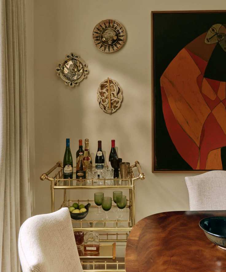 an image of the corner of a dining room painted in warm neutral tones with a brass bar cart, three decorated wall plates and large abstract piece of art