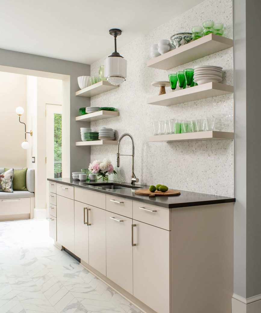 A neutral kitchen with white walls, wood cabinets, black countertops, and white floating wall shelves decorated with green glass decor and tableware