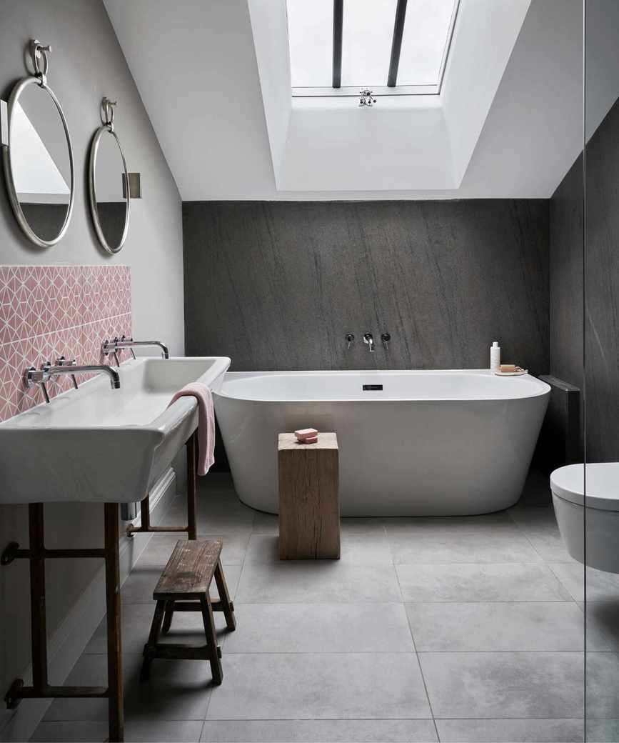 A neutral attic bathroom space with a white ceiling, wooden walls, white bathtub, wooden stools, white sink, red and white patterned backsplash and two circular mirrors above.