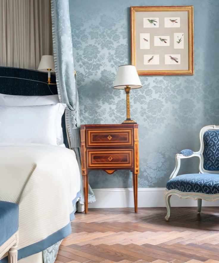 A blue wallpapered bedroom with a wooden parquet floor, with a blue armchair, framed picture, wooden bedside table and lamp, and made bed is partially pictured to the right.