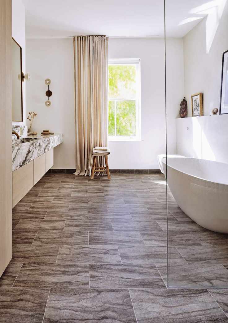 modern bathroom with white tub, glass shower wall, marble topped vanity, stool, cream drapes, vinyl floor