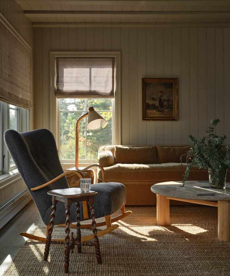 a vintage-styled paneled lakeside cottage living room with a beige velvet couch, vintage stone coffee table and a blue rocking chair