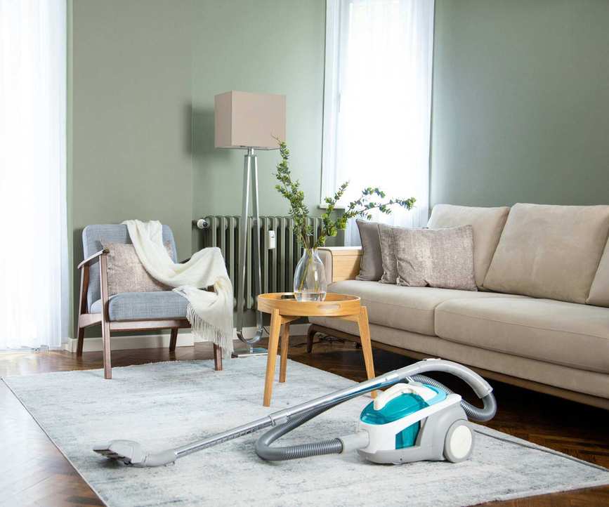 A blue and white vacuum cleaner on a cream living room rug, beside a cream couch and a small yellow side table