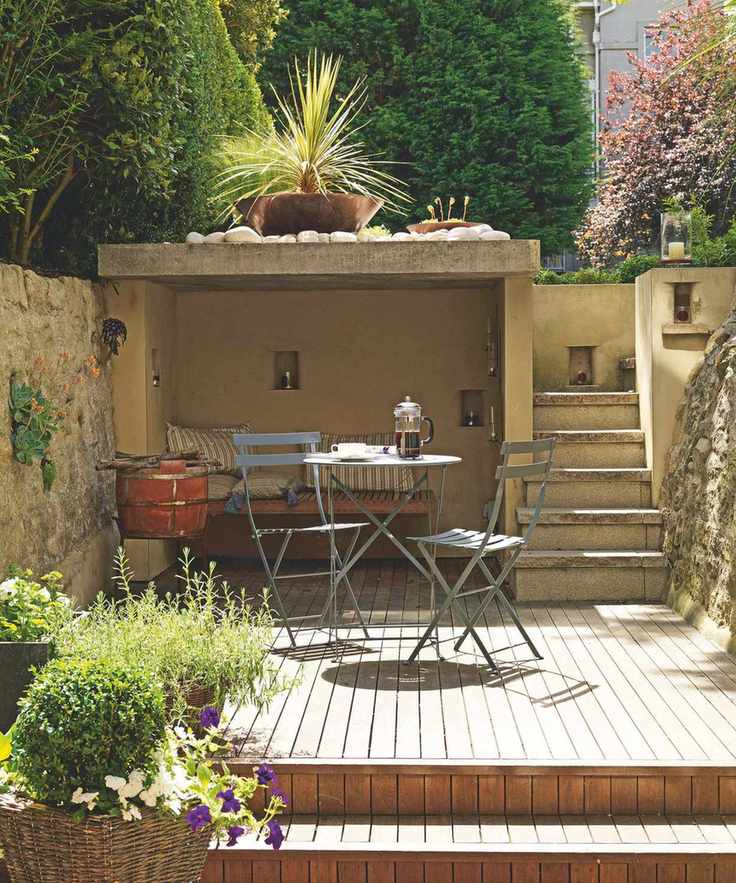 A sunny wooden outdoor decking area with metal furniture, a bricked and cushioned seating area with plants and flowers and a paved staircase
