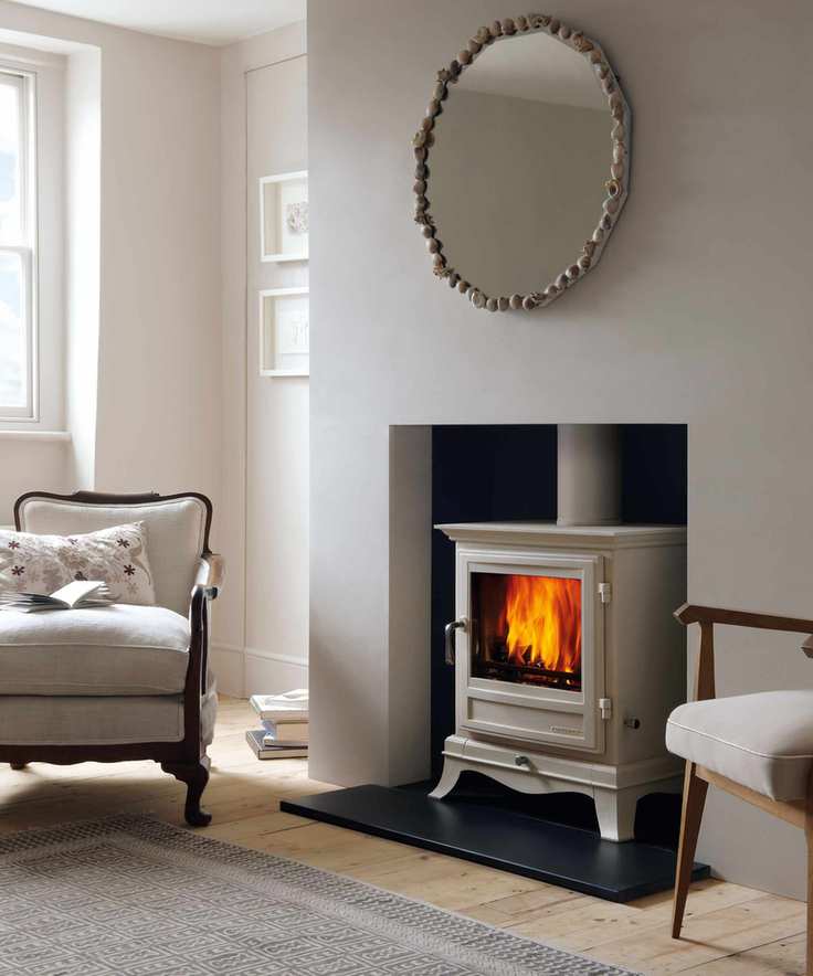 Neutral living room painted in cream, fireplace with wood burner in matching color to the room, round mirror above fireplace, light wood flooring with rug, upholstered lounge chair with cushions