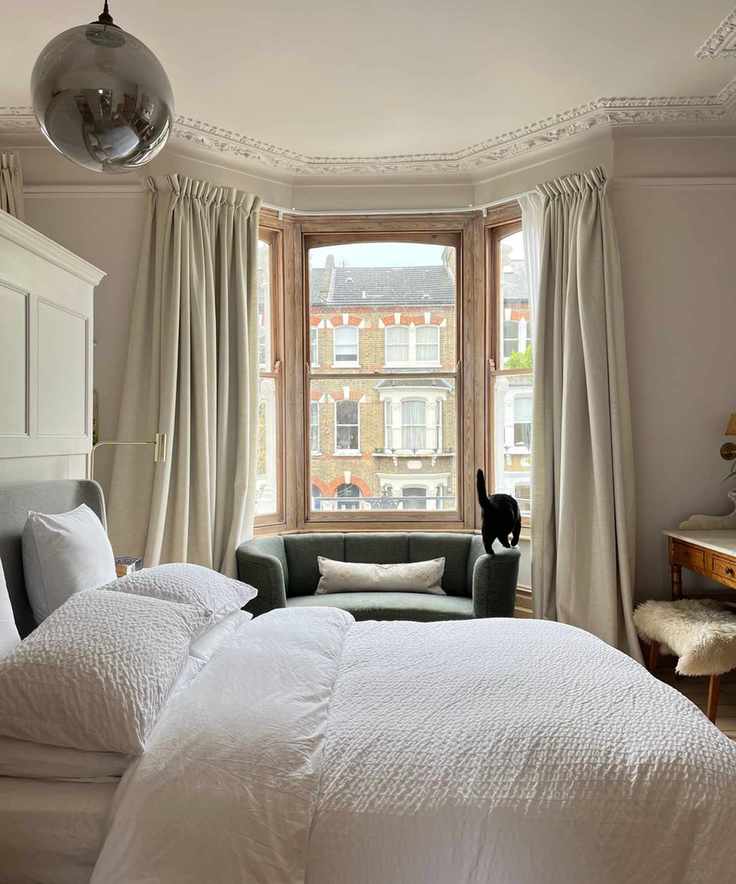 Bedroom with crown moulding, large bed with white bedding, bay window with small sofa, and wardrobe separating the space