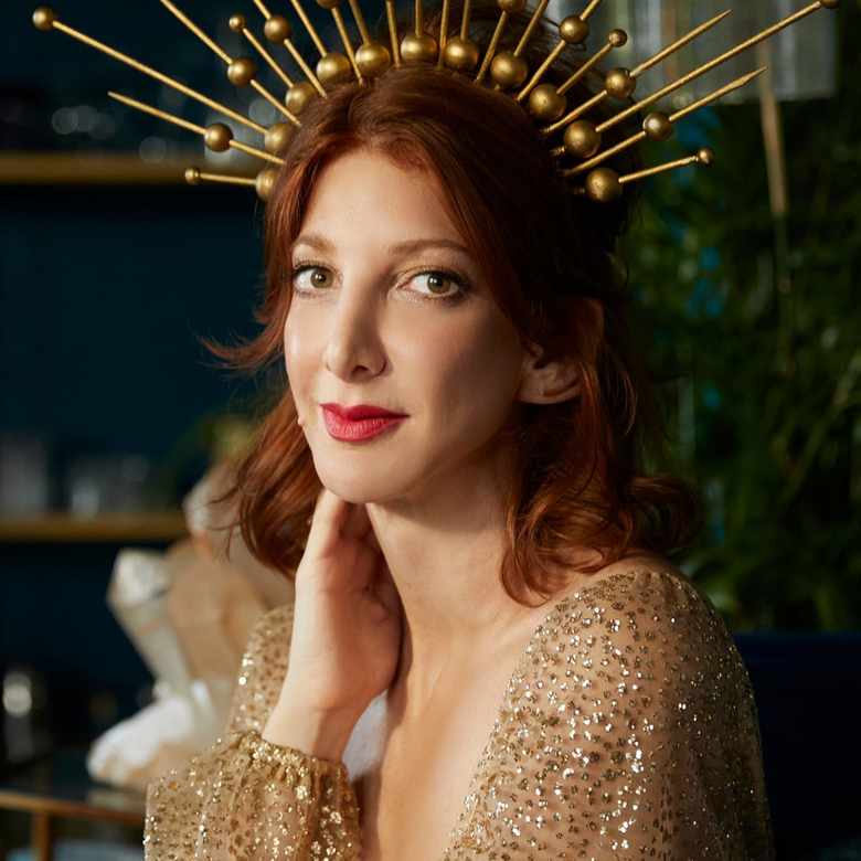 a headshot photo of astrologer Rebecca Gordon: a red haired lady wearing a gold sequin dress with a matching gold headpiece