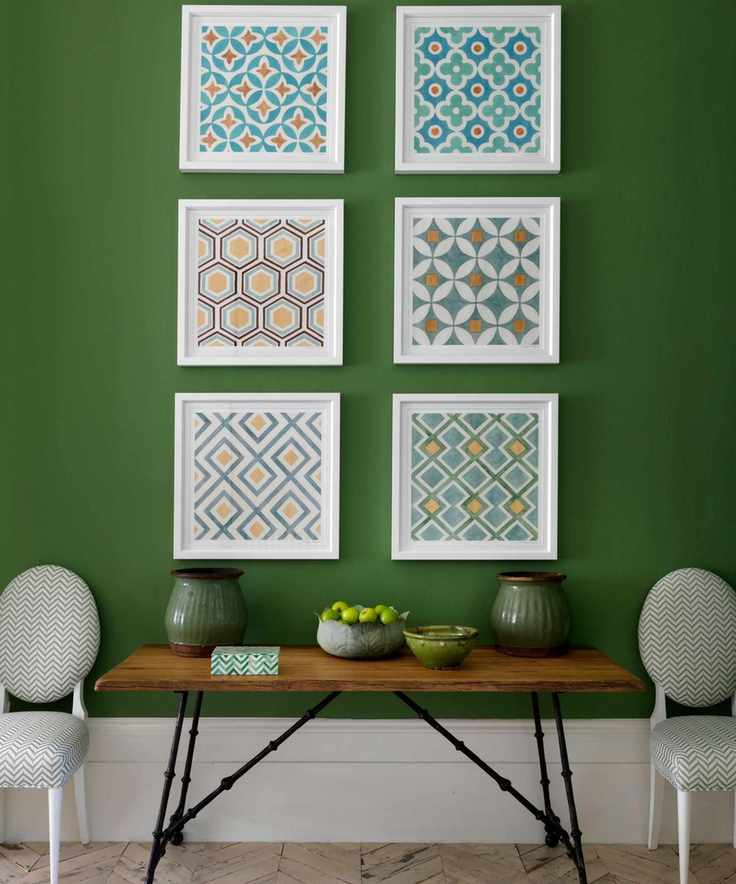 A green wall with framed abstract artwork, a wooden table with green vases and a fruitbowl, and white and green upholstered chairs.