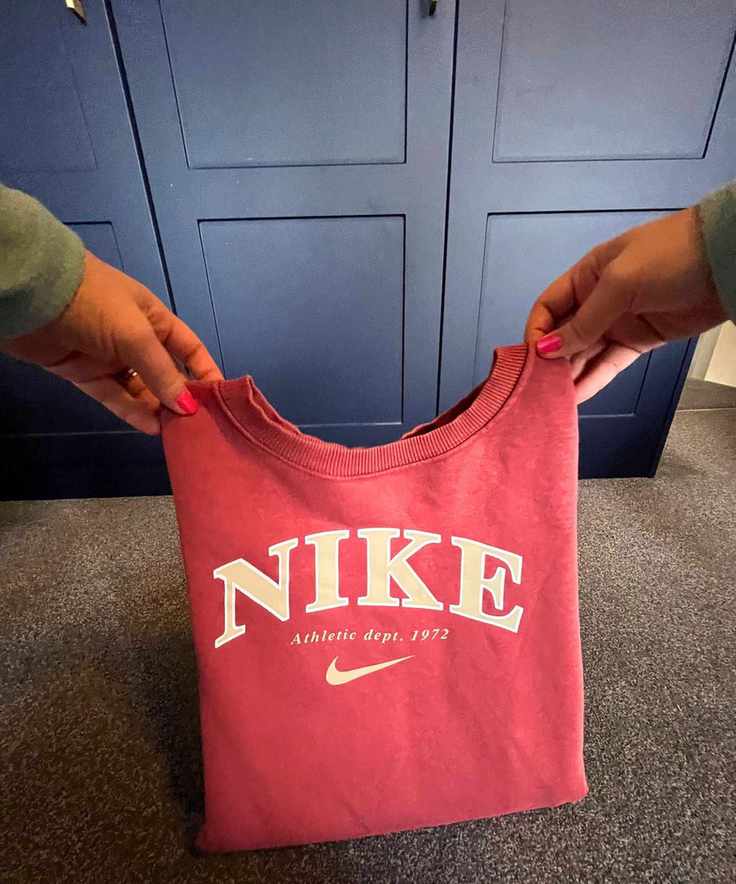 The image shows a point of view shot of a peachy-orange Nike long sleeve sweater being folded back over its bottom half on a gray carpeted floor