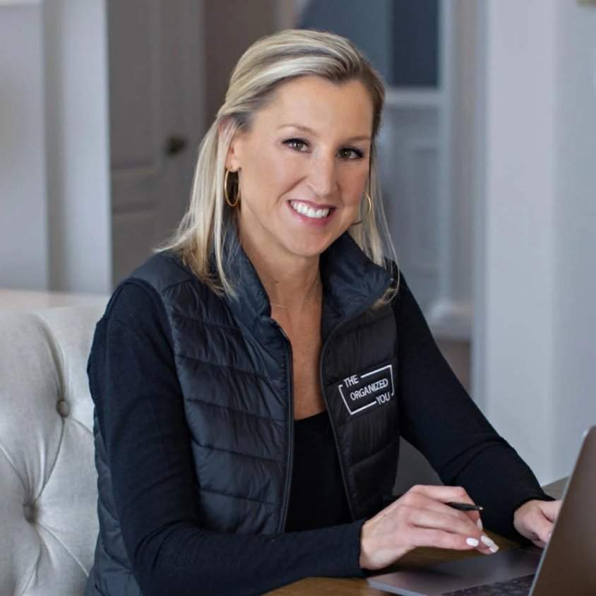 a photo of professional organizer Heather Aiello wearing all black sat behind a laptop: a blond haired lady in a long sleeved black top with a black gilet over the top with her organizing company's brand name on it