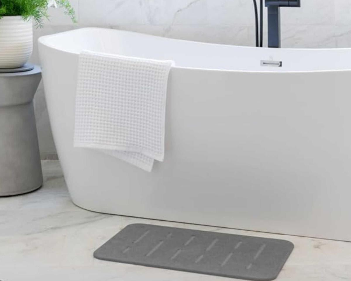 Gray stone bath mat in front of a freestanding white bathtub, and on marble veined floors.