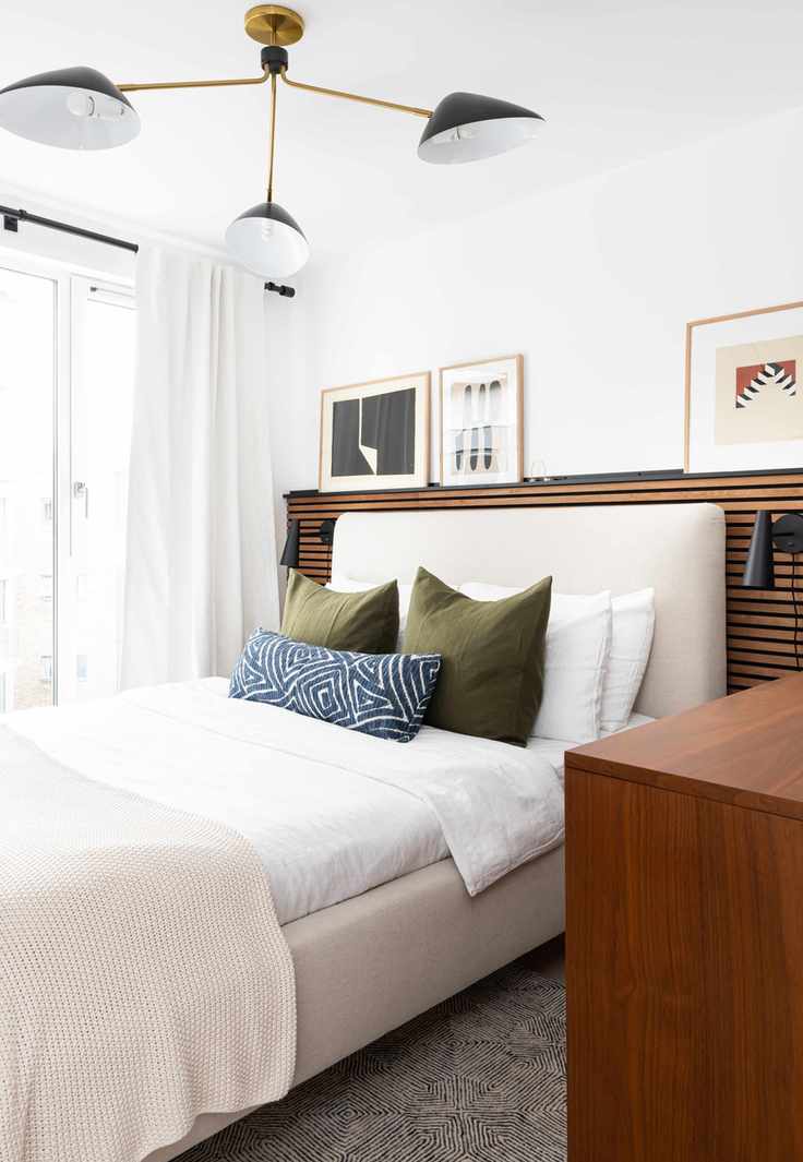 white bedroom with wood panelling behind the headboard, artwork on shelf above bed, retro pendant, white bedding, green cushions, blue cushion, wall lights, cream upholstered bed, white drapes