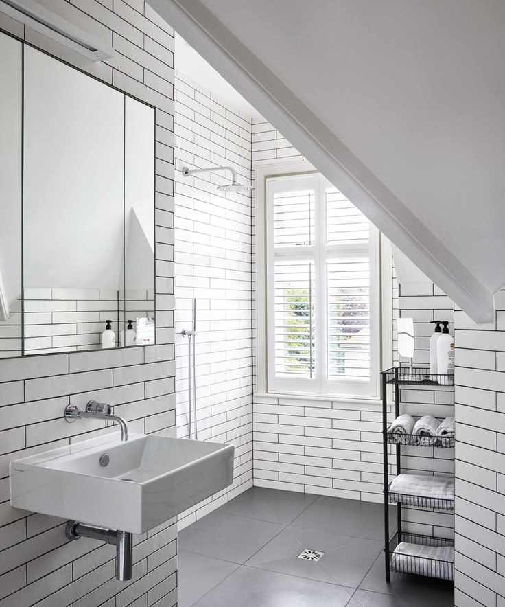 A bright white tiled attic bathroom with a white sink below a large mirror, shower, black shower caddy and large windows with white shutters.