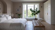 a cozy neutral bedroom with a large bed, a leather bench, a tree in front of the window, and a wall of cabinetry on the right side