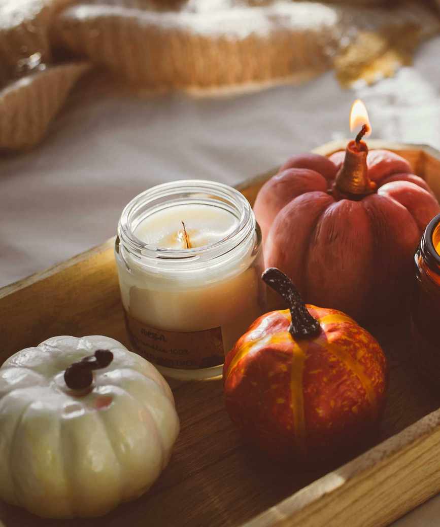 An unlit jar candle on a wooden tray next to a small pumpkin and a lit pumpkin-shaped candle