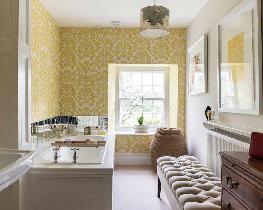 Elegant bathroom with a traditional basin and bath, showcasing an ottoman and antique chest of drawers against yellow and white wallpaper.