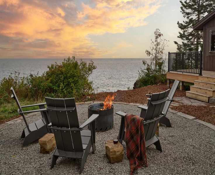 an outdoor firepit overlooking a large lake a sunset with adirondrack chairs and plaid blankets