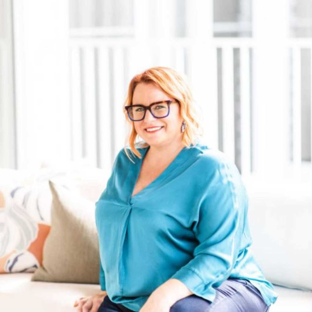 A headshot of Suzanne Butler wearing a blue shirt and large brown glasses.