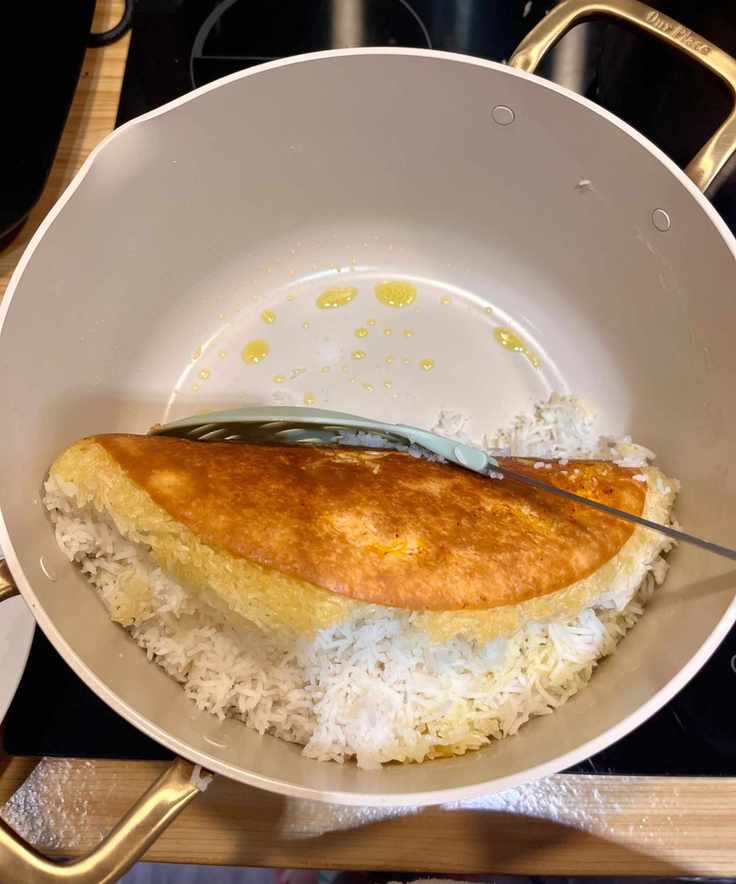 The tahdig is perfectly golden brown all over with no sticking or burning, seen here being lifted off the base of the stock pot in one piece using a large round slatted silicone utensil