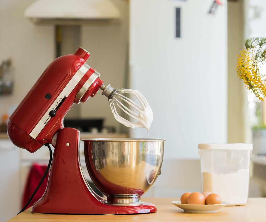 A red stand mixer on a countertop with white frosting on the whisk attachment, next to a plate of eggs and a container of flour