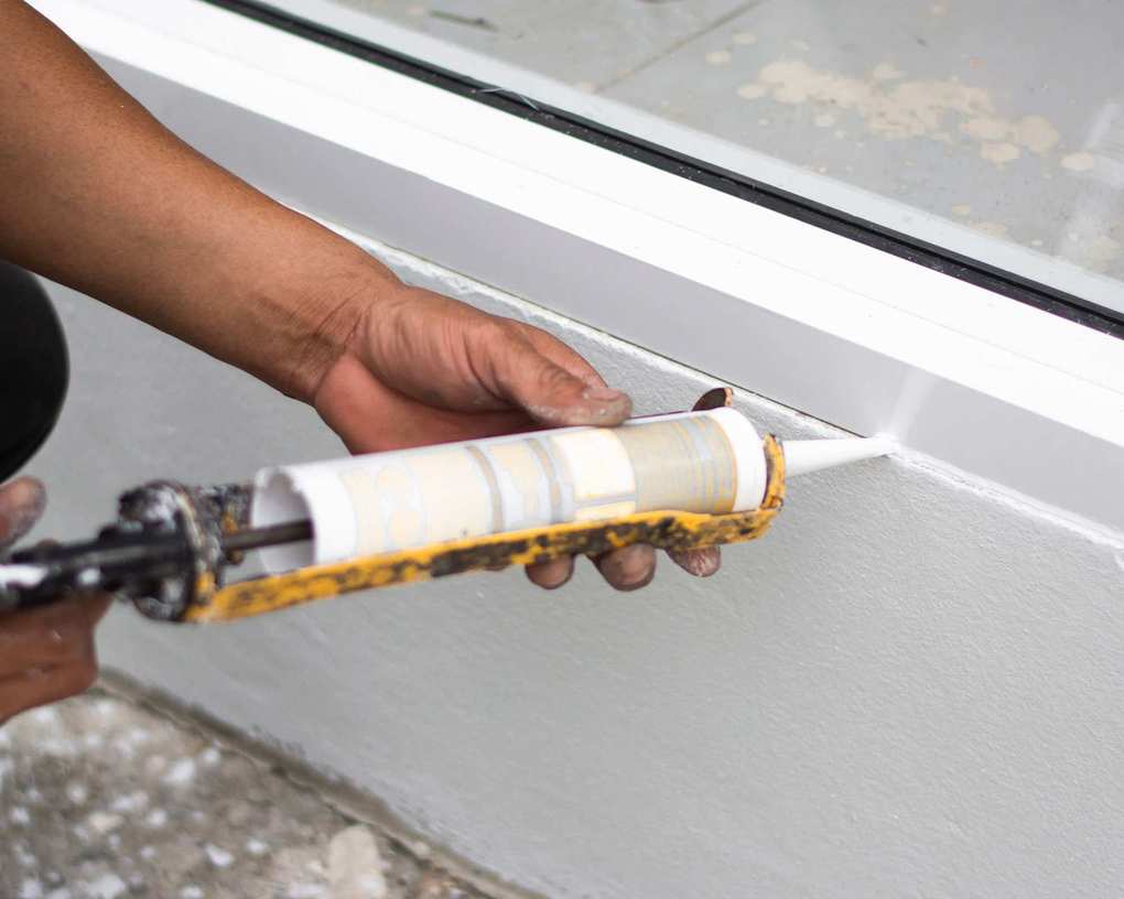 A man's hands holding a yellow caulking gun and applying caulk to the bottom of a white window frame
