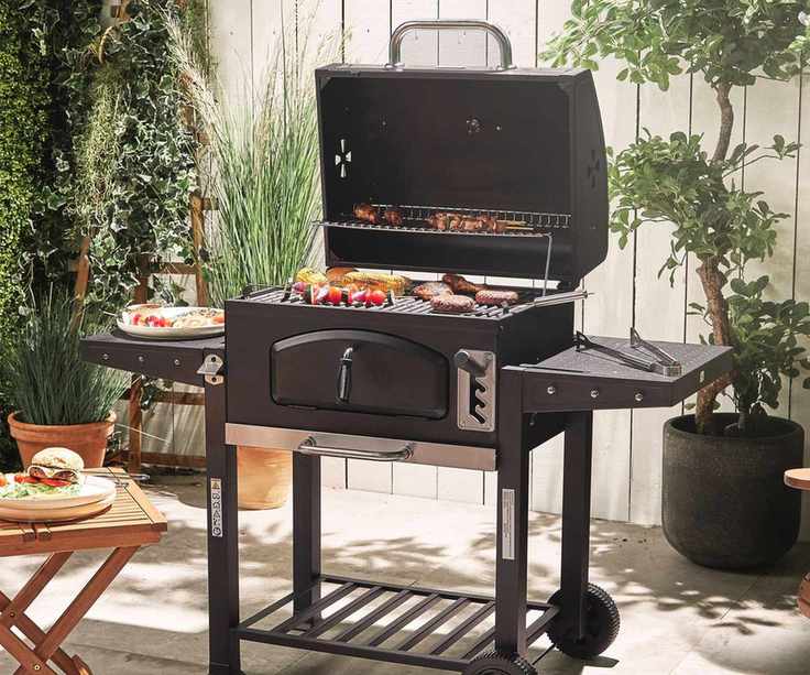 A black charcoal grill in a back garden, against a white fence, frying food
