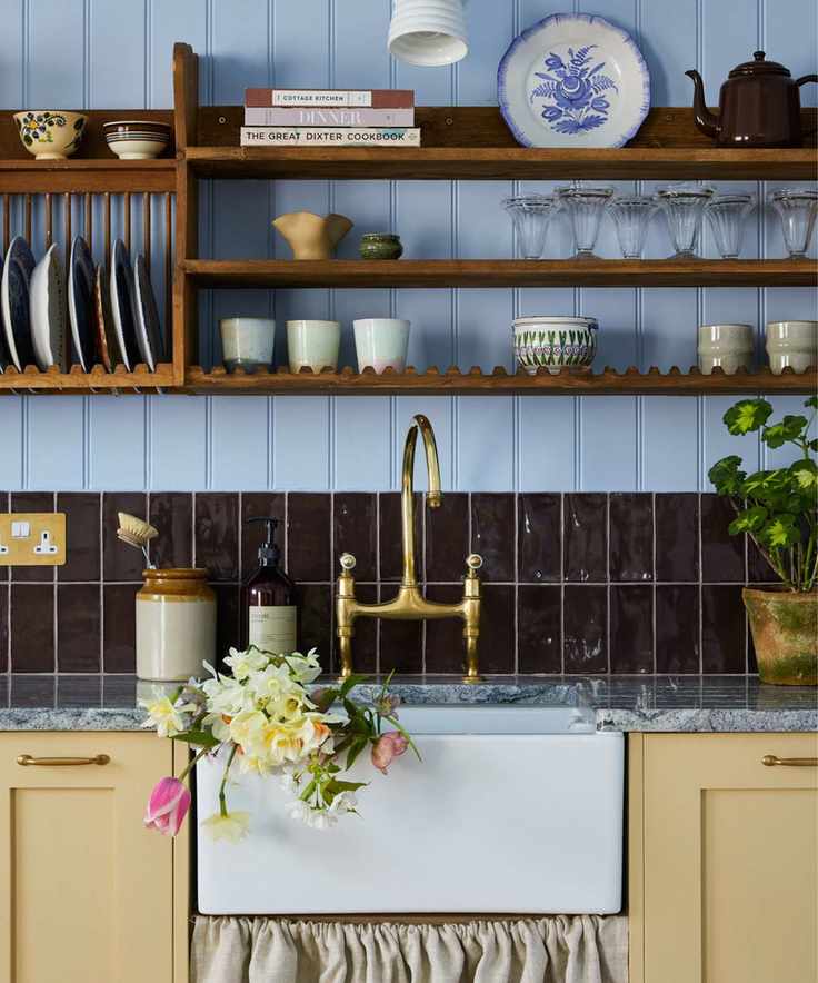Blue paneled wood wall in kitchen with wood open shelving filled with vintage ceramics and butter yellow cabinets with ceramic sink and gold tpa