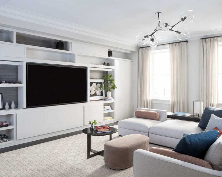 Bright living room painted white with grey sofa, large white storage unit that houses TV, cream rug and curtains, glass pendant light