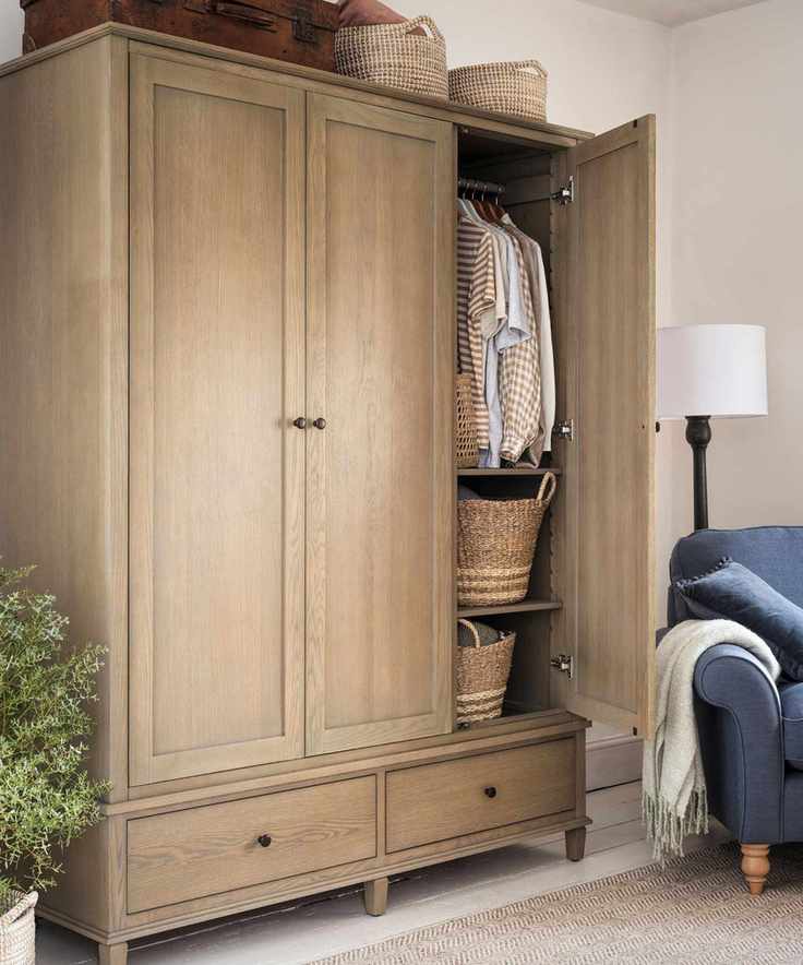 A large three-door wooden closet, with one door open showing some hanging shirts and two woven baskets. Baskets are stored on top of the wardrobe with a blue lounge chair and lamp to the right and a green potted plant to the left.