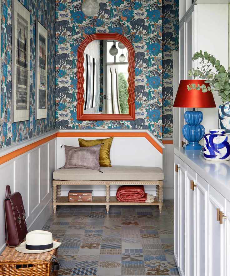 maximalist entryway with modern blue stylised wallpaper, white panelling with orange stripe, bench seat, orange mirror, artwork, patterned floor tiles, hamper, lamp, vase, seat pillows