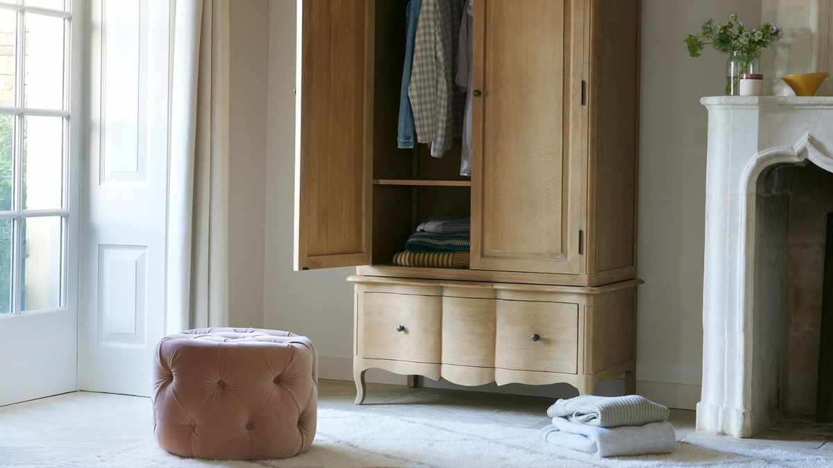 Loaf closet half open with a pink pouffe seat in front of it and a small pile of folded clothes on the floor beside it.