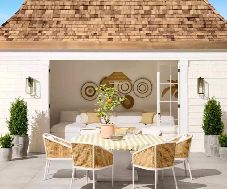 white pool house with a boho coastal style with a rectangle table and white and cane wicker chairs styled with a yellow striped tablecloth