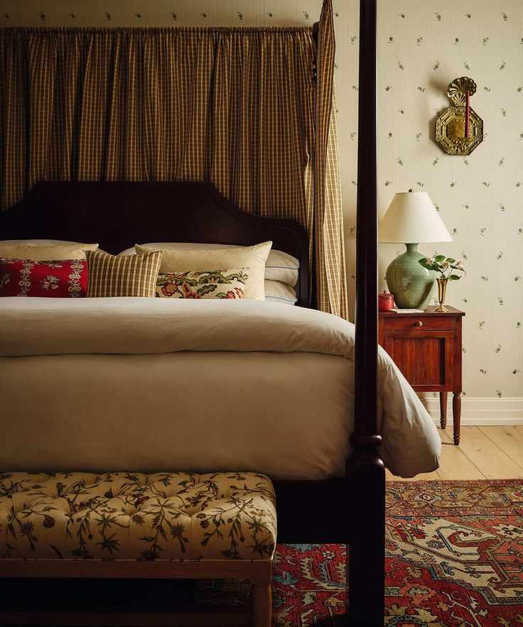 a layered primary bedroom with ditsy floral wallpaper, a dark wooden four poster bed with plaid drapes, a red vintage rug and antique furniture