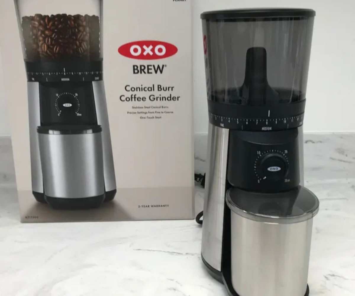 A coffee grinder with a stainless steel base and catch cup, with a black clear plastic hopper on top, in front of its branded OXO box, on a white marble counter top.
