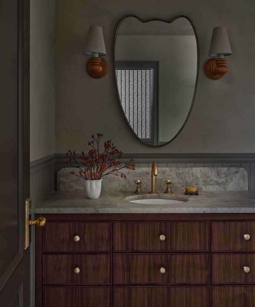 a cottage bathroom vanity area with dark brown wooden vanity, stone counter and a curved mirror with two wooden sconces either side