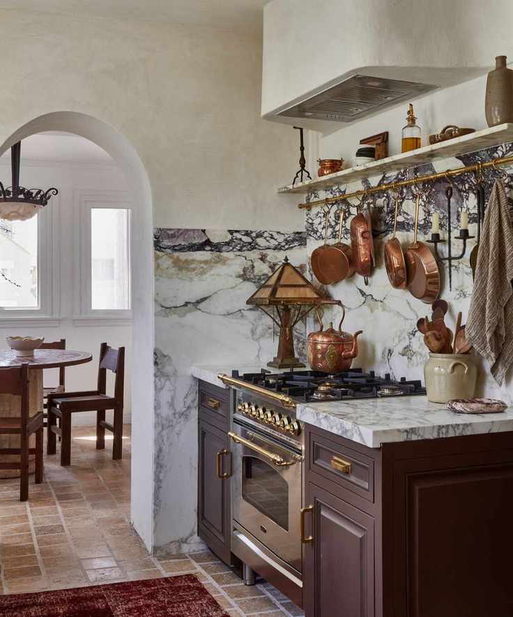 A Vintage kitchen with brown cabinets, a marble countersplash, a brass pot rack on the wall, and a range cooker topped with a vintage copper teapot