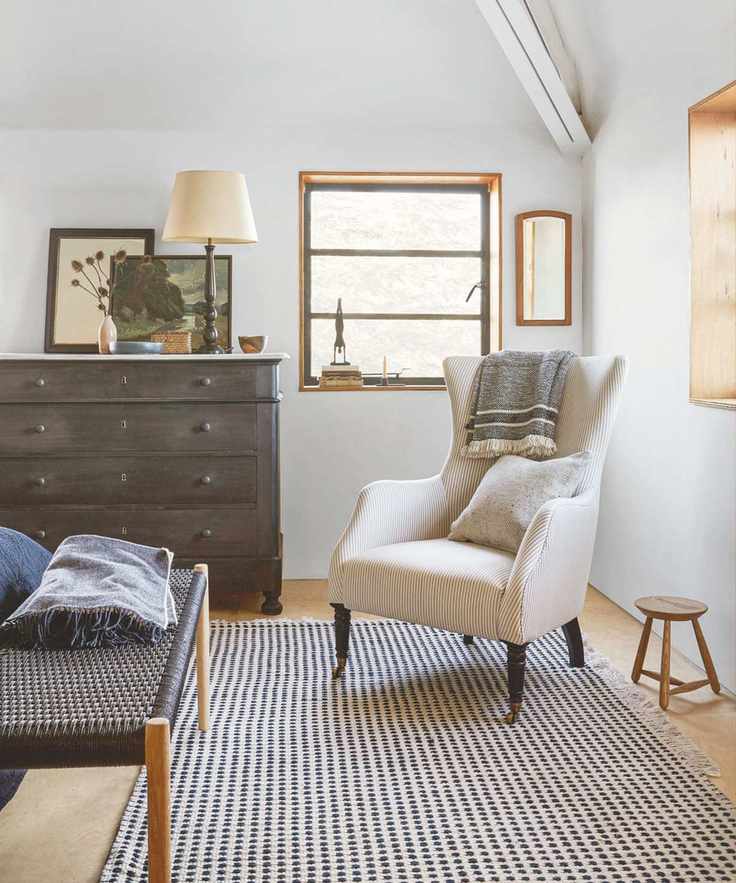 A neutral bedroom space with a black and white rug, white upholstered chair next to a small wooden stool, brown dresser with lamp and artworks and brown bench.