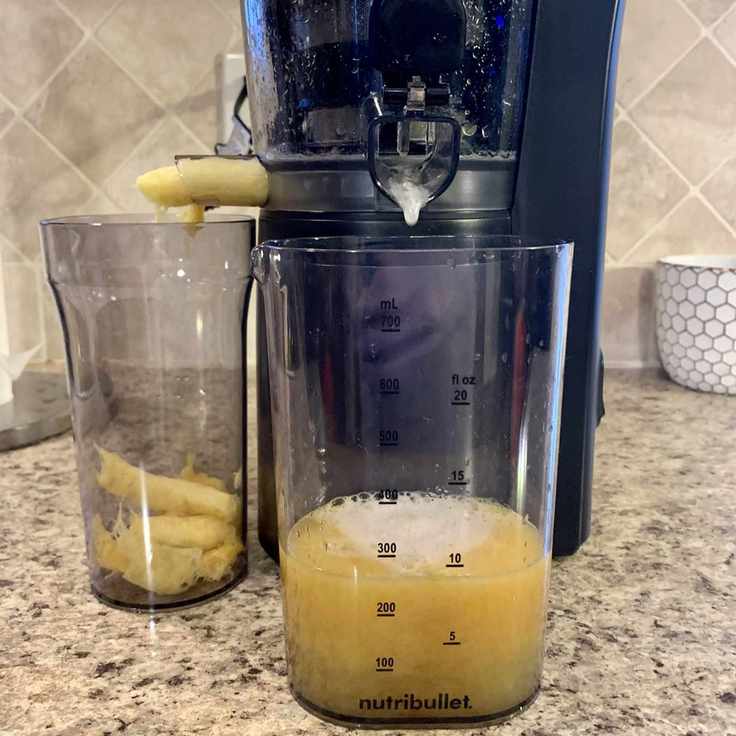A close up of the base of a black plastic cold-press juicer, dispensing pale yellow fruit solids into a beaker on its left, and pale yellow juice into a clear beaker in front of it on a granite kitchen countertop.