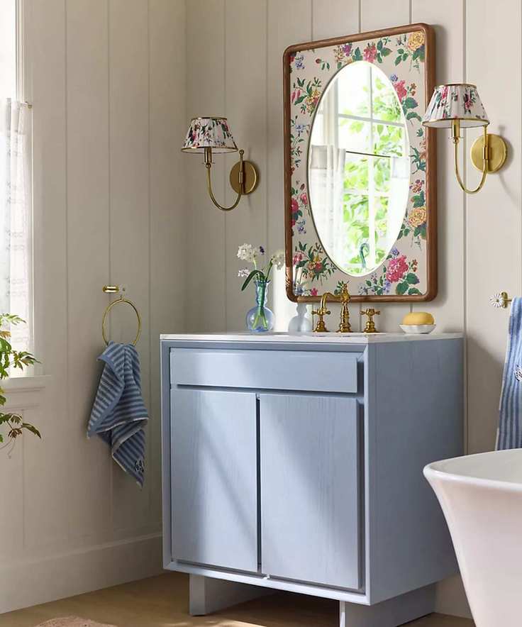 A small bathroom with cream paneled walls, a blue vanity, brass hardware, and a floral mirror