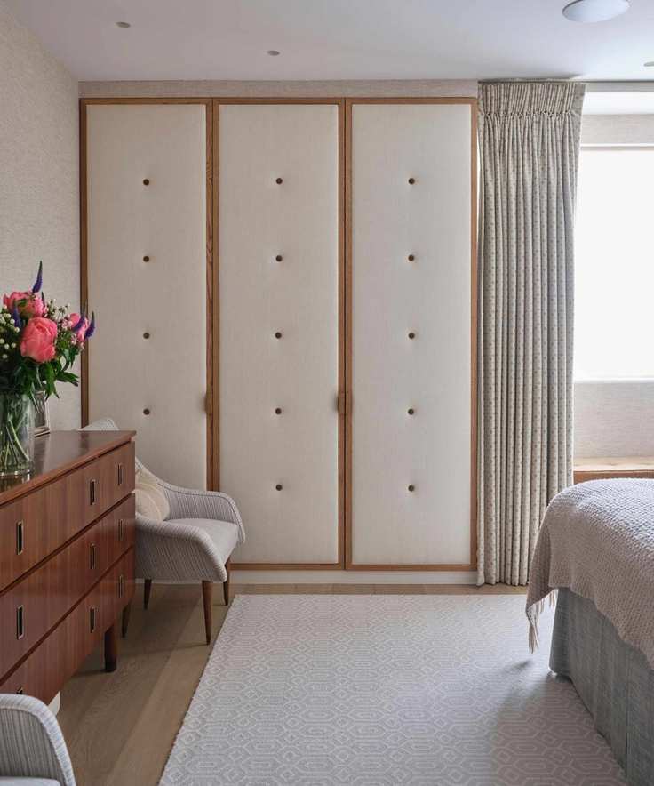 A cream and wood padded closet in a bedroom with a cream rug, wooden sideboard with vase of bright pink flowers, and grey chair.