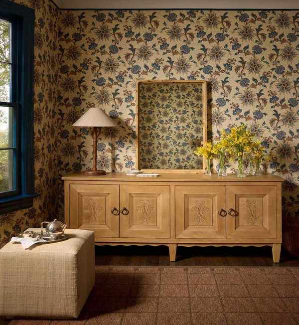 a room with a checker printed rug, a light wood credenza, a fabric ottoman, a mirror leaning on the credenza, a lamp, and an arrangement of flowers, all in a room with warm, floral wallpaper