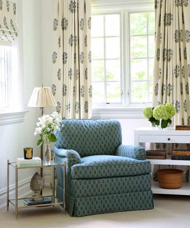 Cozy corner of living room with cream patterned curtains, matching blind, blue armchair, side tables with flowers, books and ornaments
