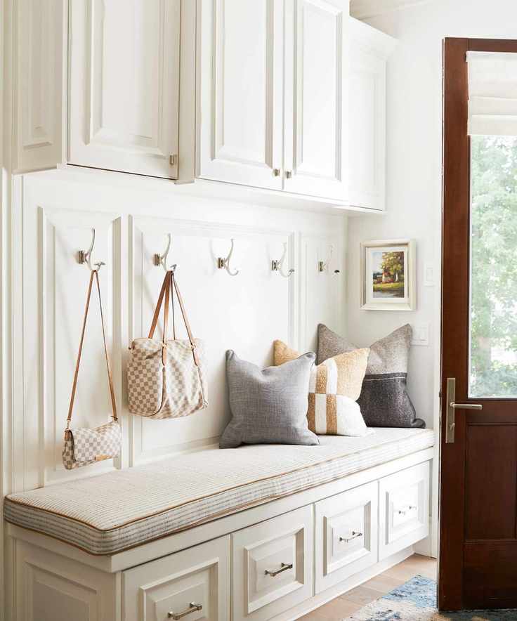 A light cream painted storage bench with a matching cushion on top in an entryway. Bags hanging from hooks on the wall.